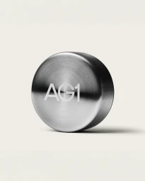 Close-up of a circular brushed stainless steel AG1 container lid casting a soft shadow on a plain neutral background.