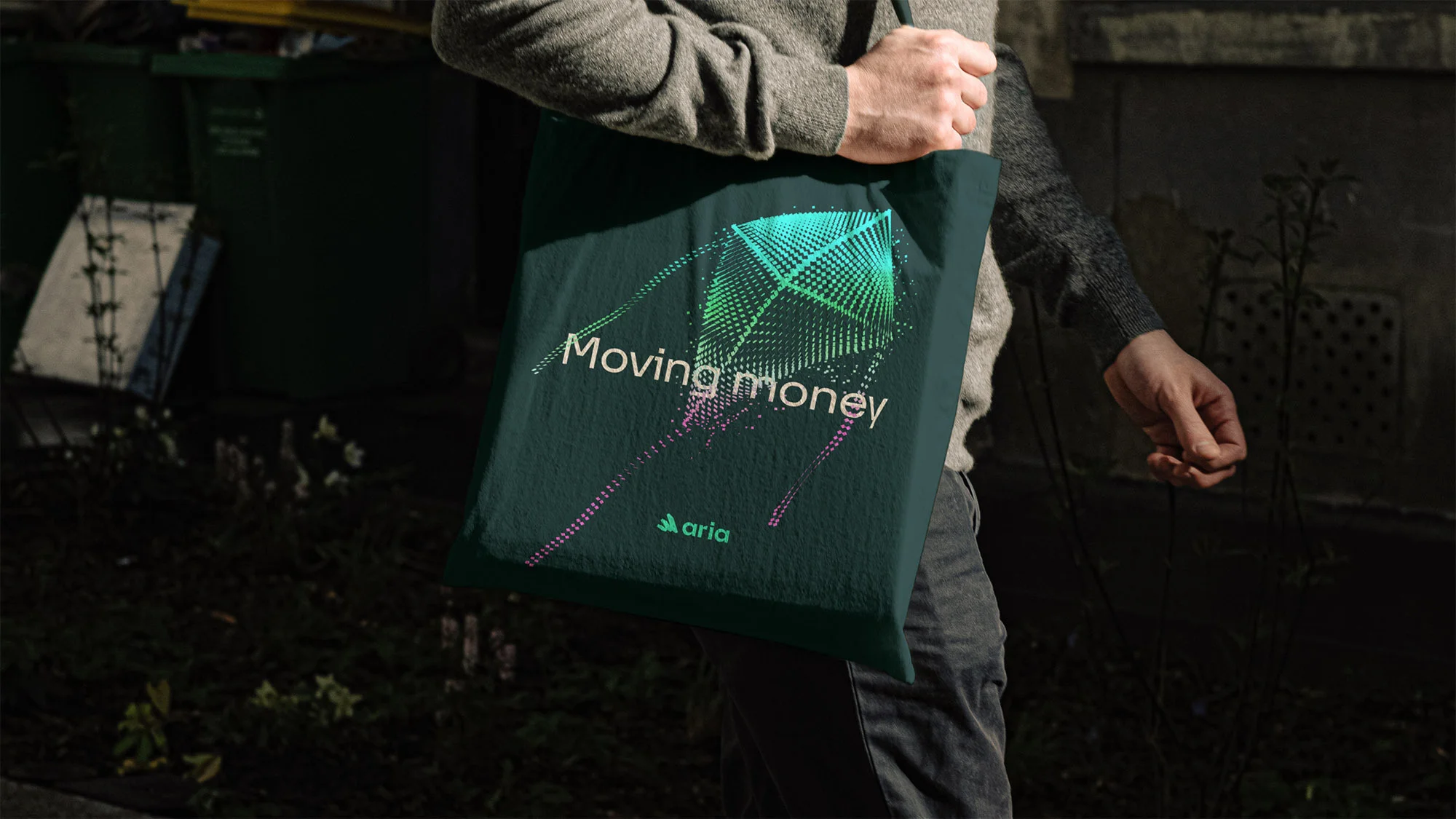 A person wearing a gray sweater and jeans carrying a green tote bag with a geometric design and the words 'Moving money' and 'Aria'.