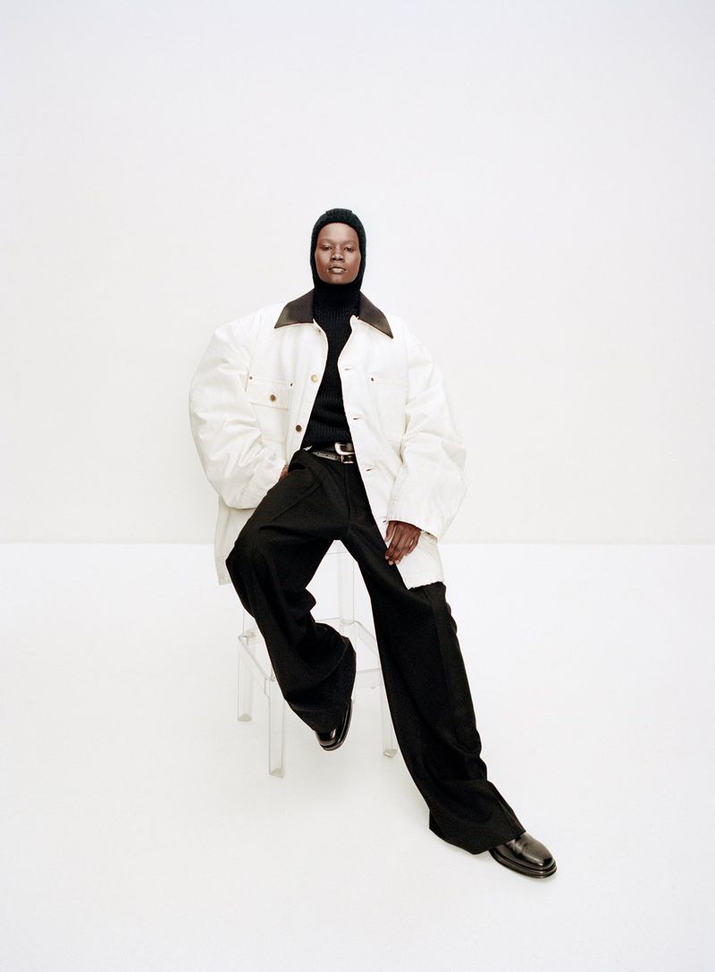 A Black woman in an off-white jacket, black top and pants, and black shoes sits on a transparent chair against a white background.