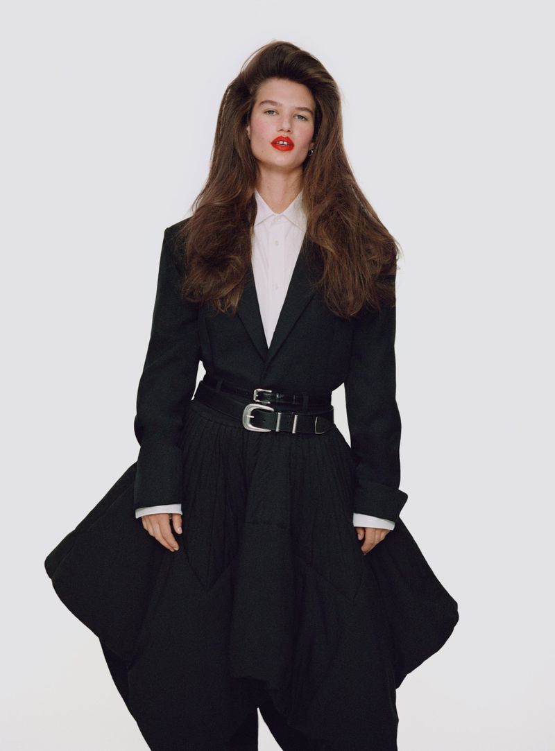A single adult female fashion model stands in a photo studio, wearing a black oversized blazer, voluminous skirt, and bold red lipstick.