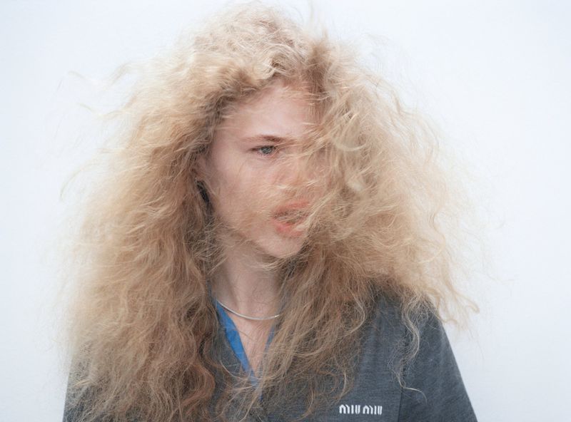 Portrait of a blonde adult female model with long, flowing, frizzy hair, wearing a casual grey top in a bright, minimalist setting.