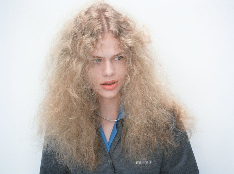Close-up studio shot of a blond adult female model wearing a gray cardigan and a blue shirt posing against a white background.