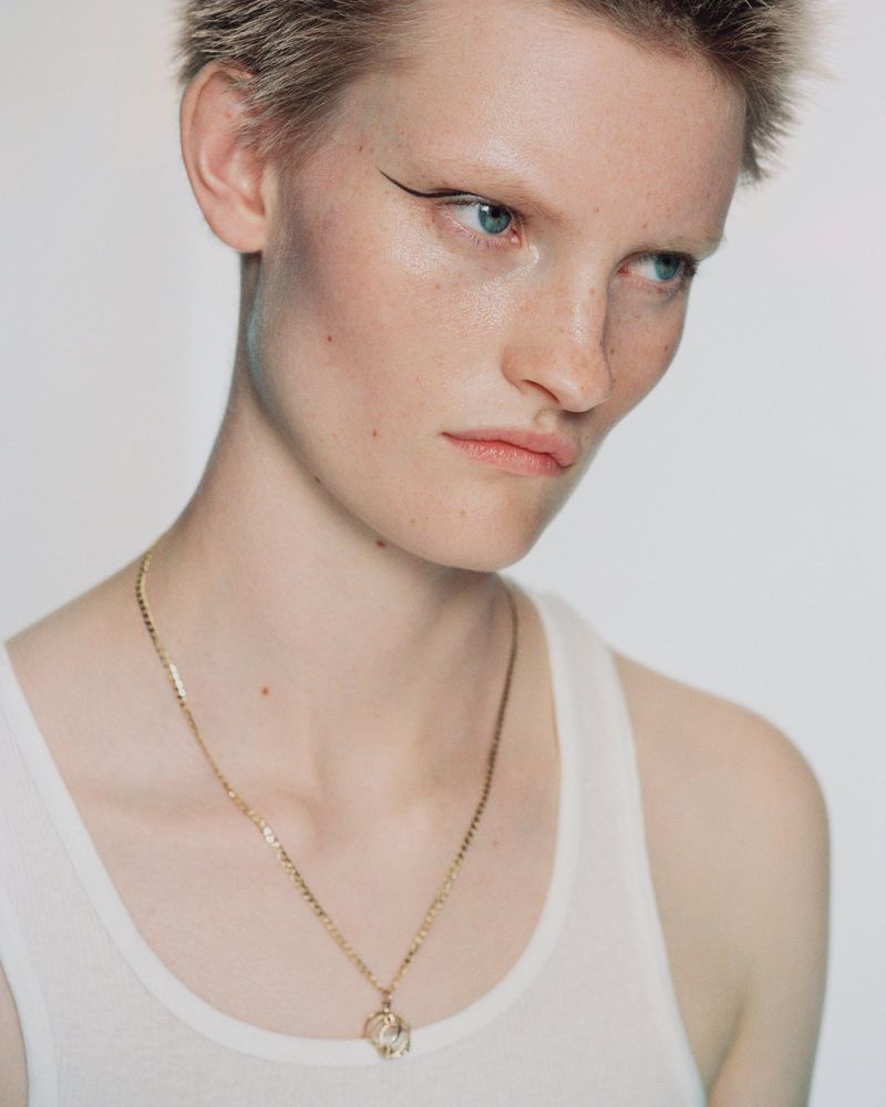 Close-up of a young adult female with blonde pixie cut, wearing a white tank top and gold necklace against a white background.