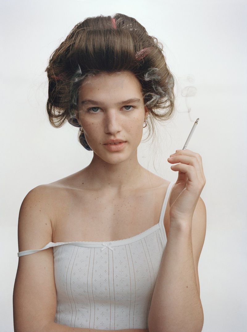 Portrait of a young fair-skinned woman with hair rollers smoking a cigarette against a white backdrop, wearing a white tank top.
