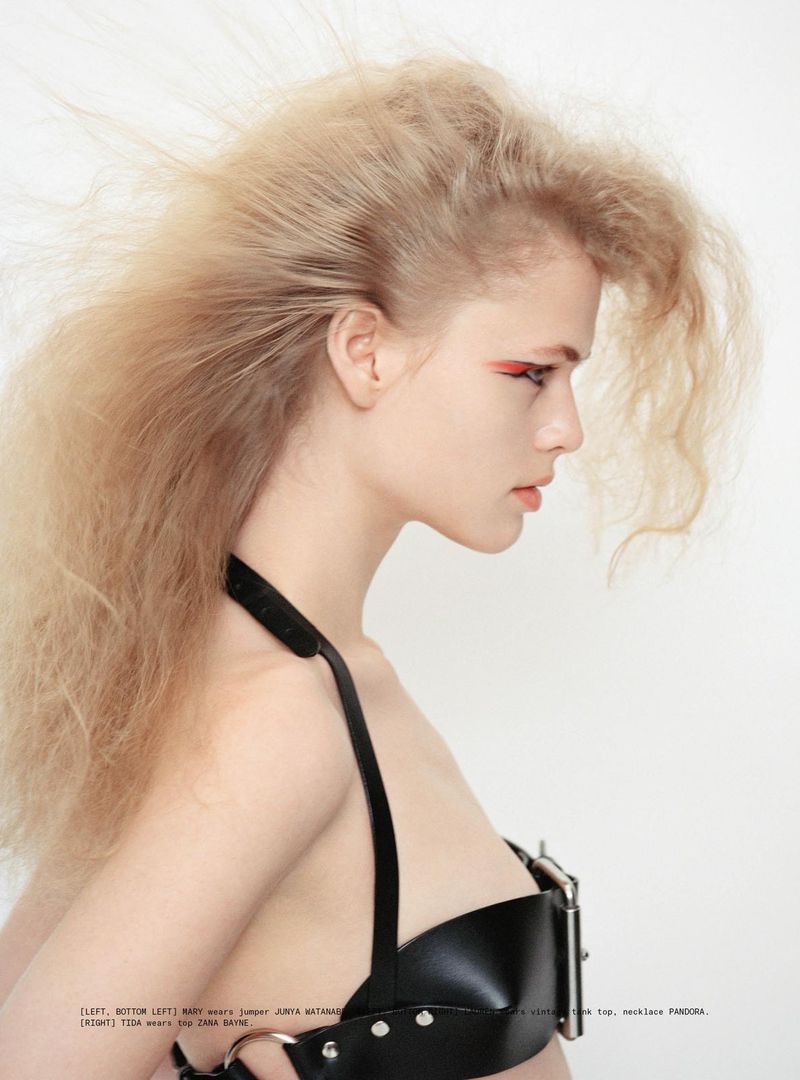 Portrait of a young woman with blond hair and a black leather harness top, set against a white background in a studio.
