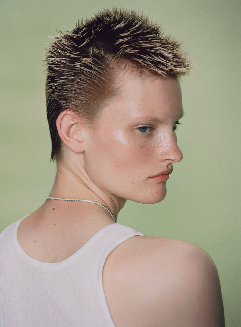 Close-up portrait of a person with a short, textured hairstyle, fair skin, and a white tank top against a light green backdrop.