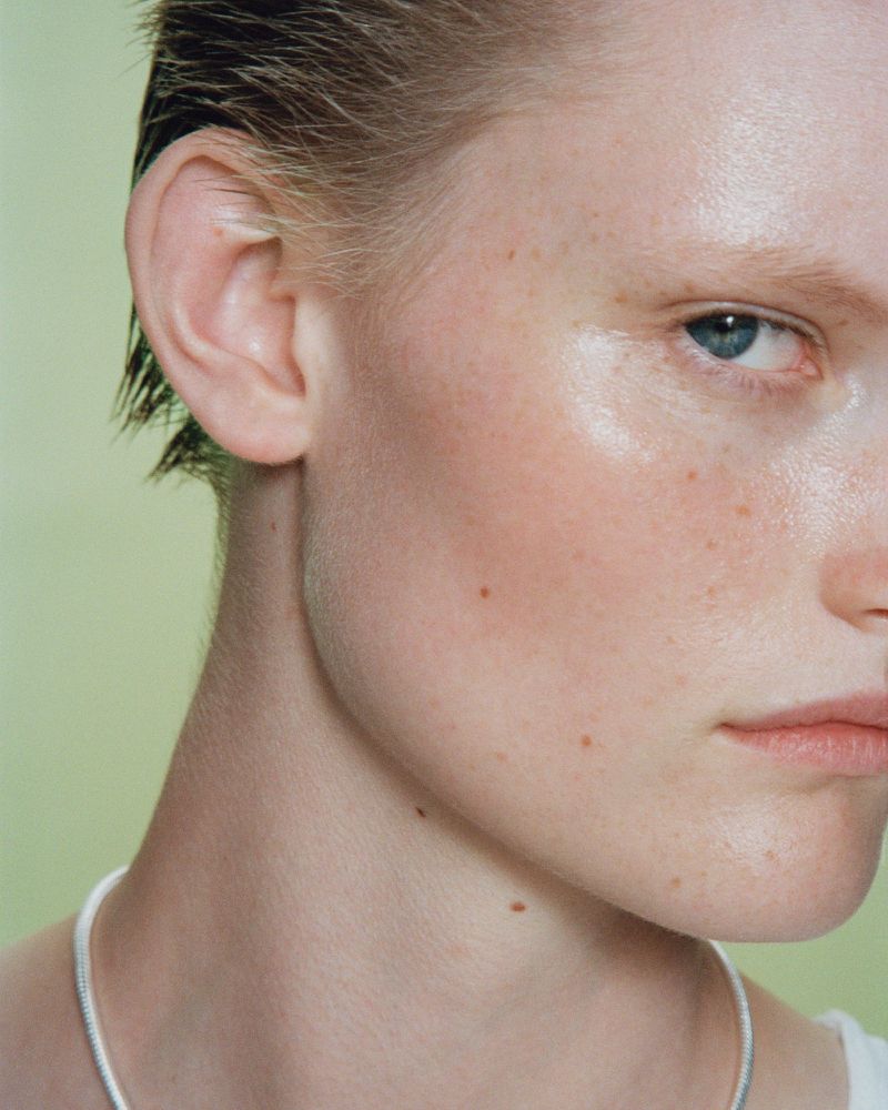 Close-up studio portrait of an adult female with short, wet hair, freckles, and fair skin, wearing a delicate silver necklace against a pale green background.