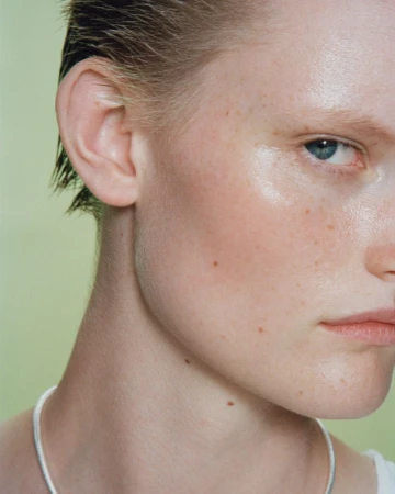 Close-up studio portrait of an adult female with short, wet hair, freckles, and fair skin, wearing a delicate silver necklace against a pale green background.