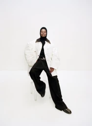 Model wearing an oversized white jacket and black trousers seated on a transparent acrylic stool against a seamless white studio background