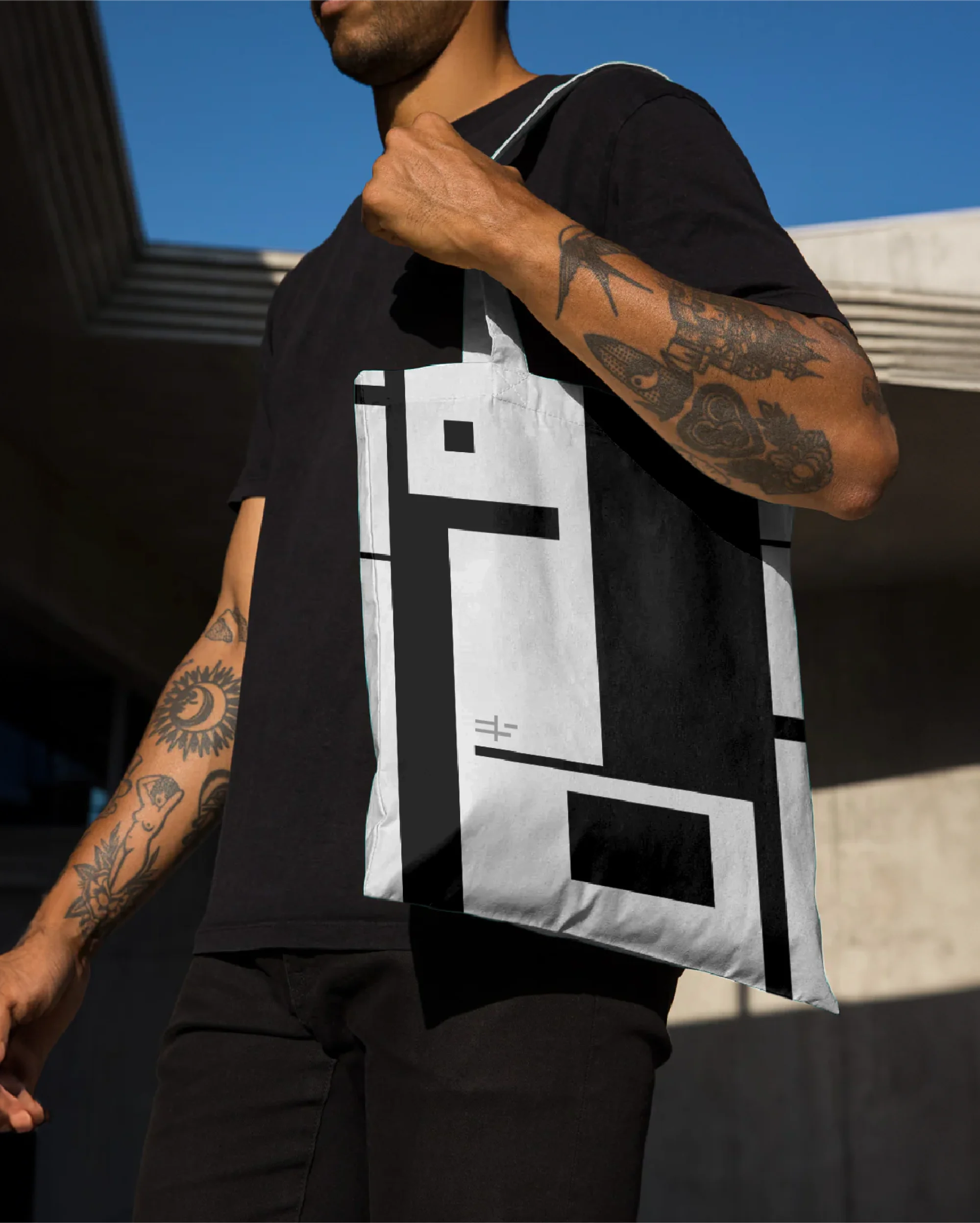 A man in a black t-shirt carries a black and white tote bag with a geometric design against a blue sky background.