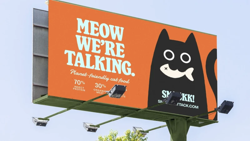 Billboard ad for SMKKK! brand cat food features a cartoon black cat head with a fish in its mouth on an orange background. 