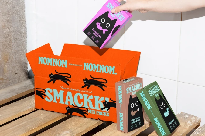 Hand placing SMACKK! cat food package into an orange box with black cat illustrations; several packages are on a wood pallet.