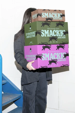Woman in oversized black suit carries stack of three Smackk cat food boxes in pink, green, and brown on white tiled background.
