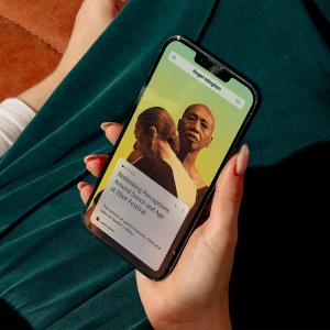 Close-up of hands holding a smartphone displaying a green-themed mobile website with portraits and text, worn over a dark green fabric garment.