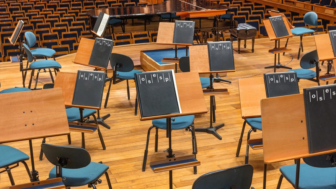 Interior shot of empty concert hall with rows of blue chairs, wooden floors, and music stands with black and tan panels.