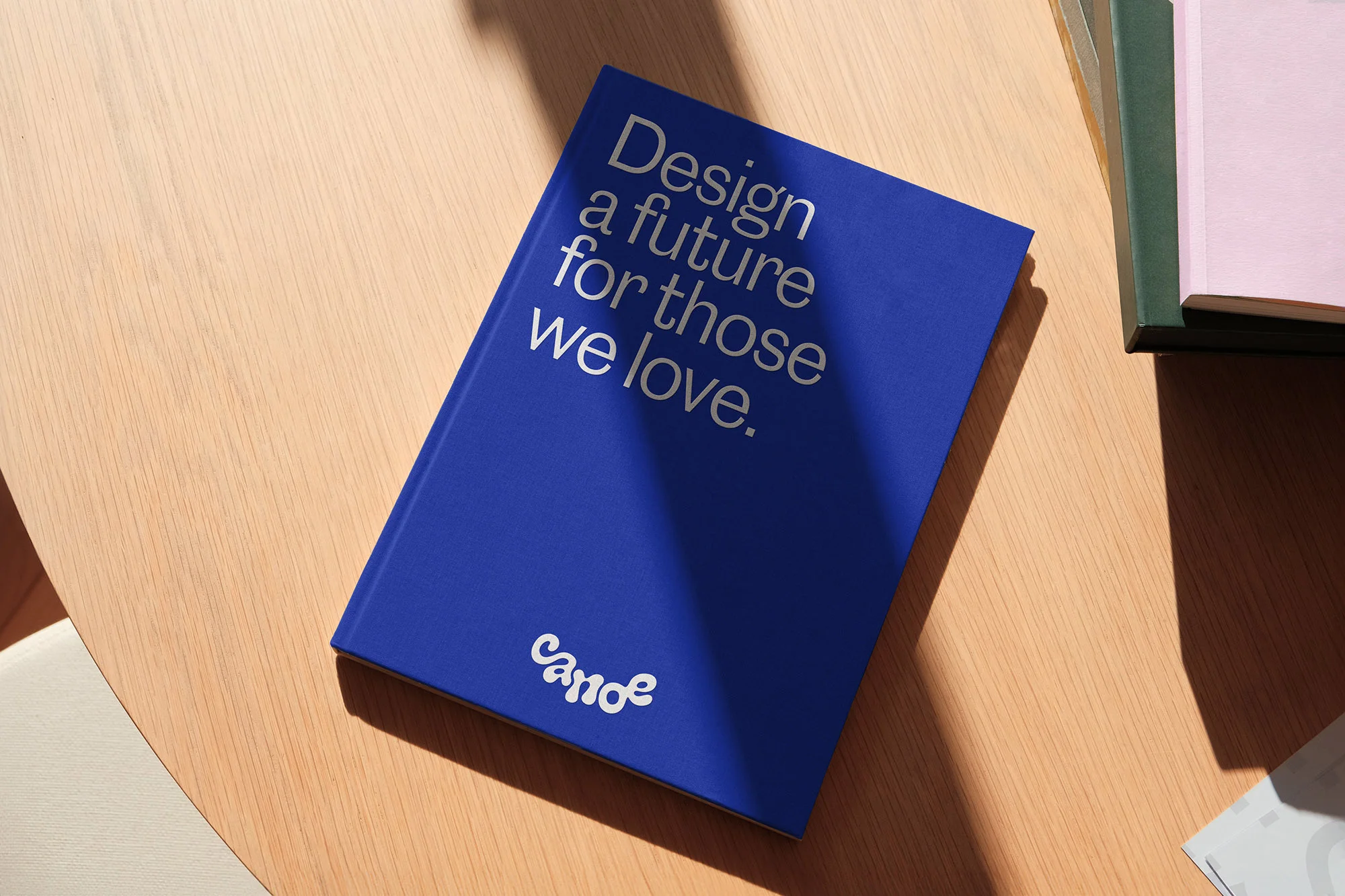 Blue hardback book, titled 'Design a future for those we love,' sits on a light wood table, bathed in dappled sunlight and shadows.