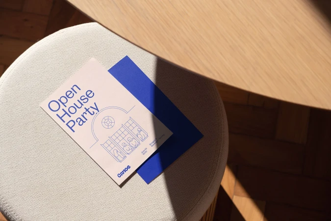 Invitation card for an Open House Party on a textured stool near a light wood table with shadows and geometric minimalist illustration.