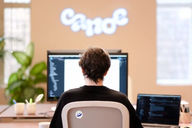 A person from behind seated at a desk with two computer screens in an office with the Canoe logo on the wall