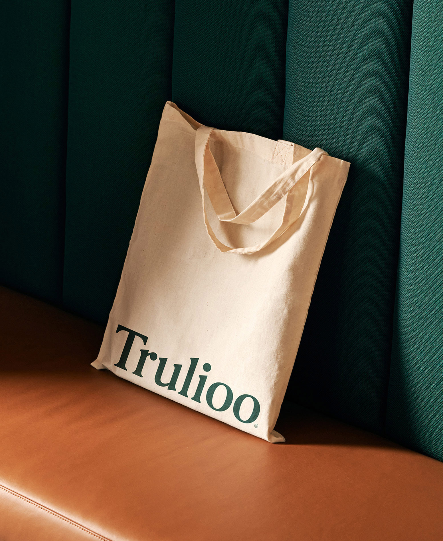 Beige canvas tote bag with dark green 'Trulioo' logo resting on a caramel-colored leather surface against a dark green textured wall.