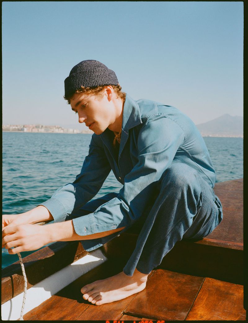 Young adult man on boat, wearing a knit beanie and a matching blue shirt and pants, with a coastal cityscape visible in background.