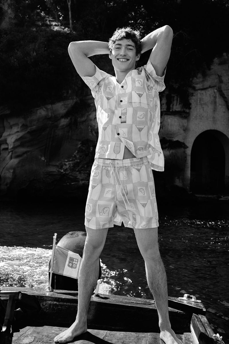 Young man in matching patterned shirt and shorts stands barefoot on a boat, posing with hands behind his head, with rocky backdrop.