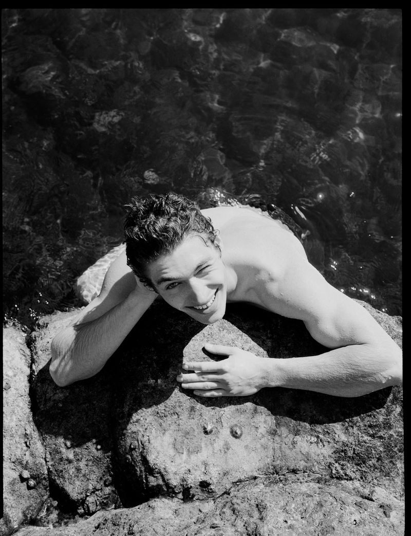 Black and white portrait of a smiling man lying on rocks in the water, looking at the camera with a relaxed facial expression.