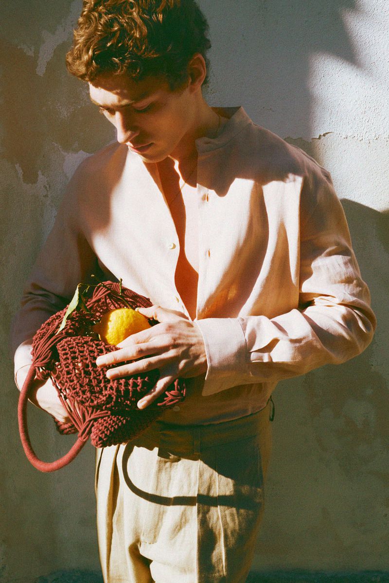 Young man in a linen shirt holds a string bag with a lemon, posing against a textured wall in warm, diffused sunlight.
