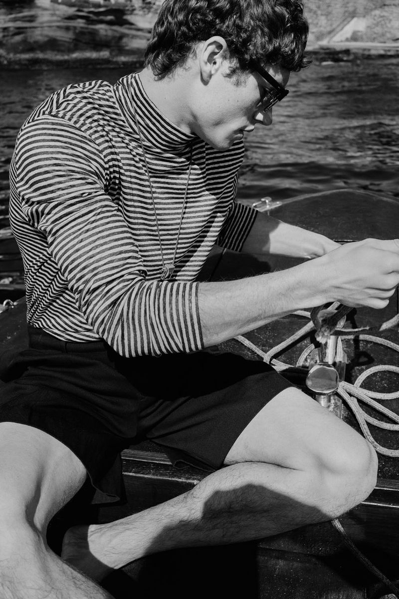 Black and white shot of a man in sunglasses wearing a striped shirt and shorts on a boat, capturing a timeless, nautical aesthetic.