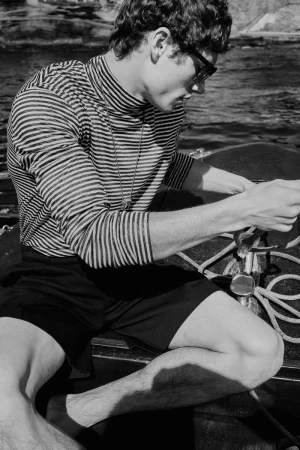 Black and white shot of a man in sunglasses wearing a striped shirt and shorts on a boat, capturing a timeless, nautical aesthetic.