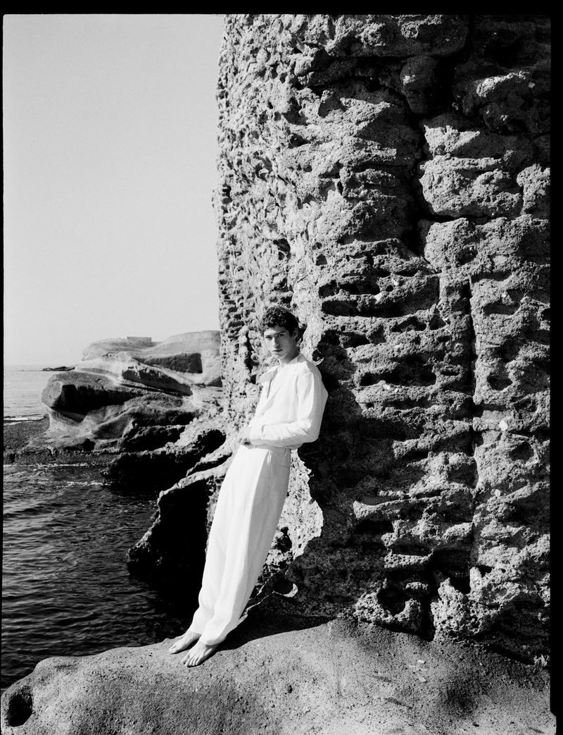 A young adult stands barefoot in white clothing, leaning against a weathered rock formation by the sea in black and white.