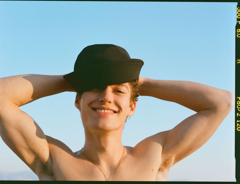 Smiling shirtless man with a hat, holding hands behind head against a blue sky backdrop, bathed in warm, natural light.