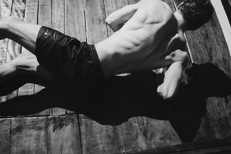 Monochrome shot of a shirtless man in shorts lying on a wooden surface, showing his back muscles and a strong shadow.