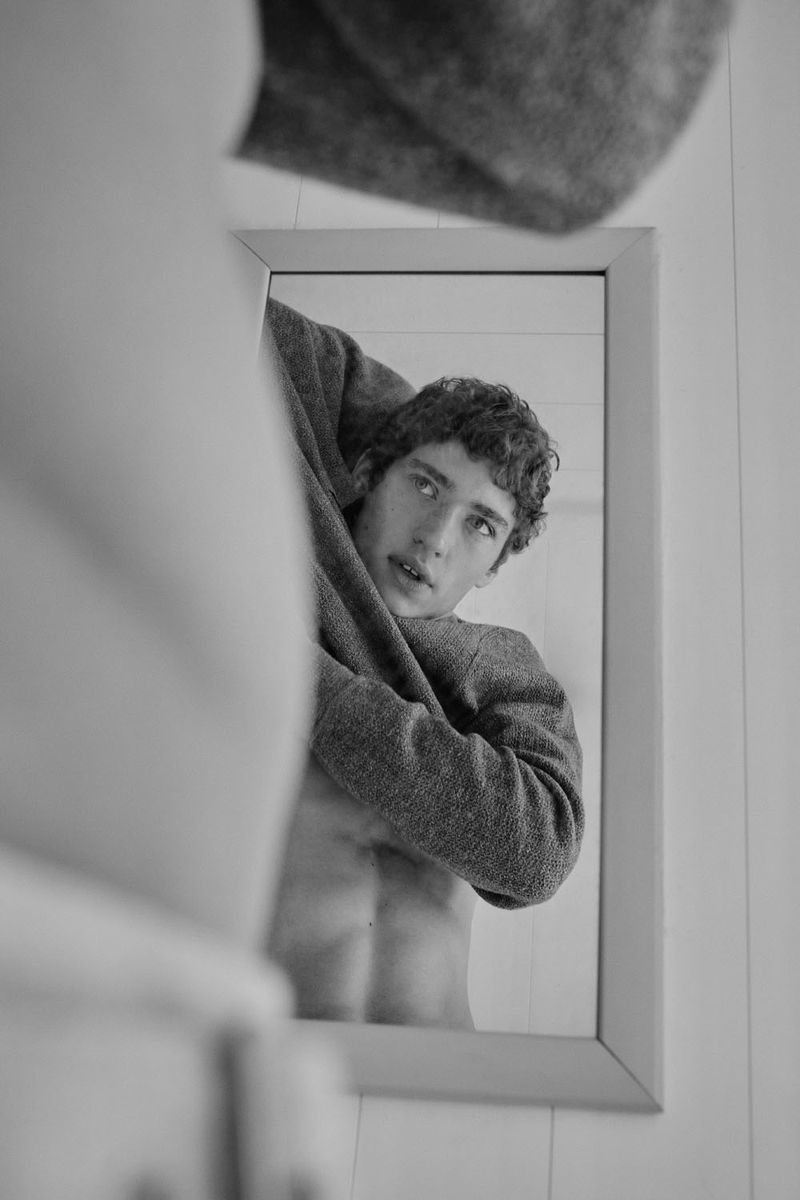 Adult male reflected in mirror, taking off grey sweater; indoor setting with white walls; black and white photography. Casual style.