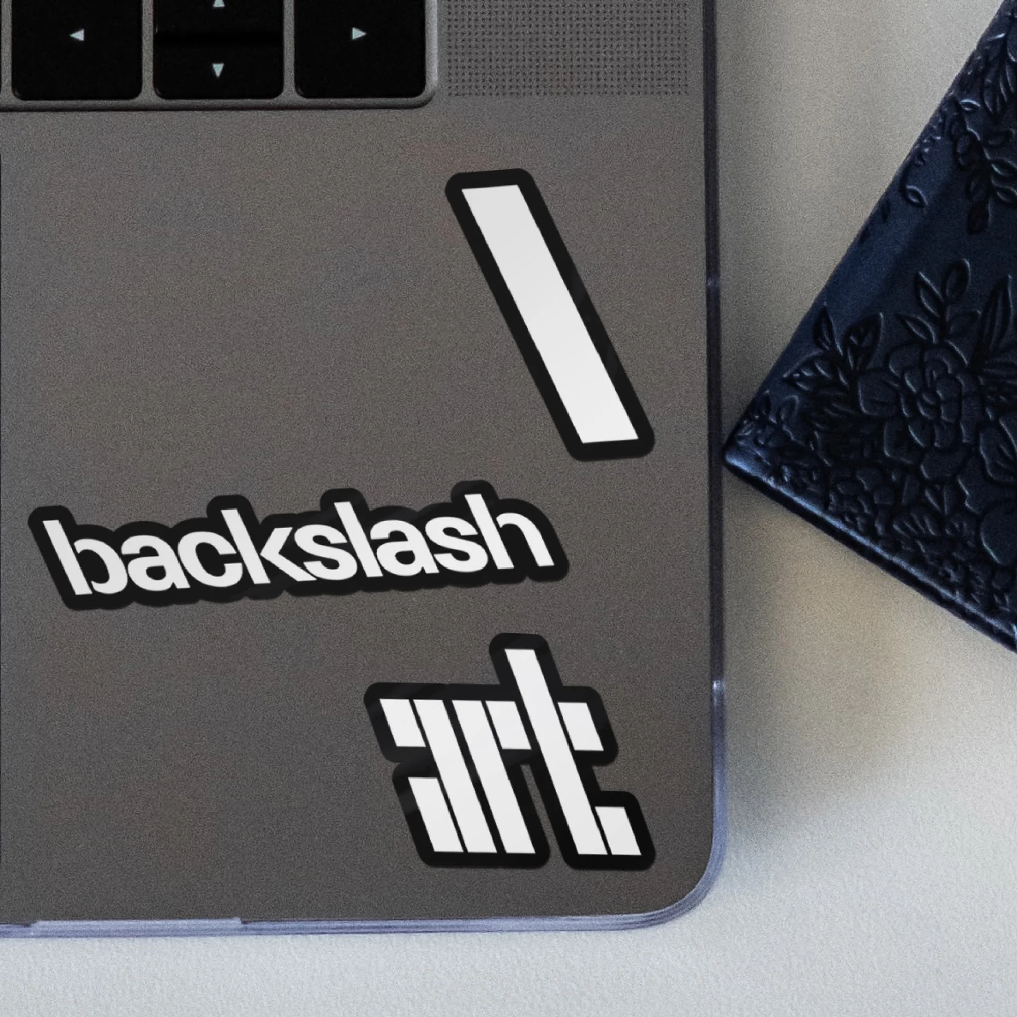 Close-up of a gray laptop surface with stickers: a line, "backslash" text, and graphic of the letters "art", next to a navy book.