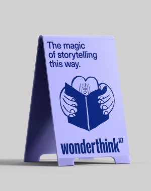 A-frame sidewalk sign in light purple with blue typography reading The magic of storytelling this way and a stylized line-art illustration.