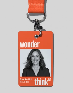 An orange lanyard and ID badge hanging against a light grey wall featuring a monochrome portrait of a woman named Brooke Hill.