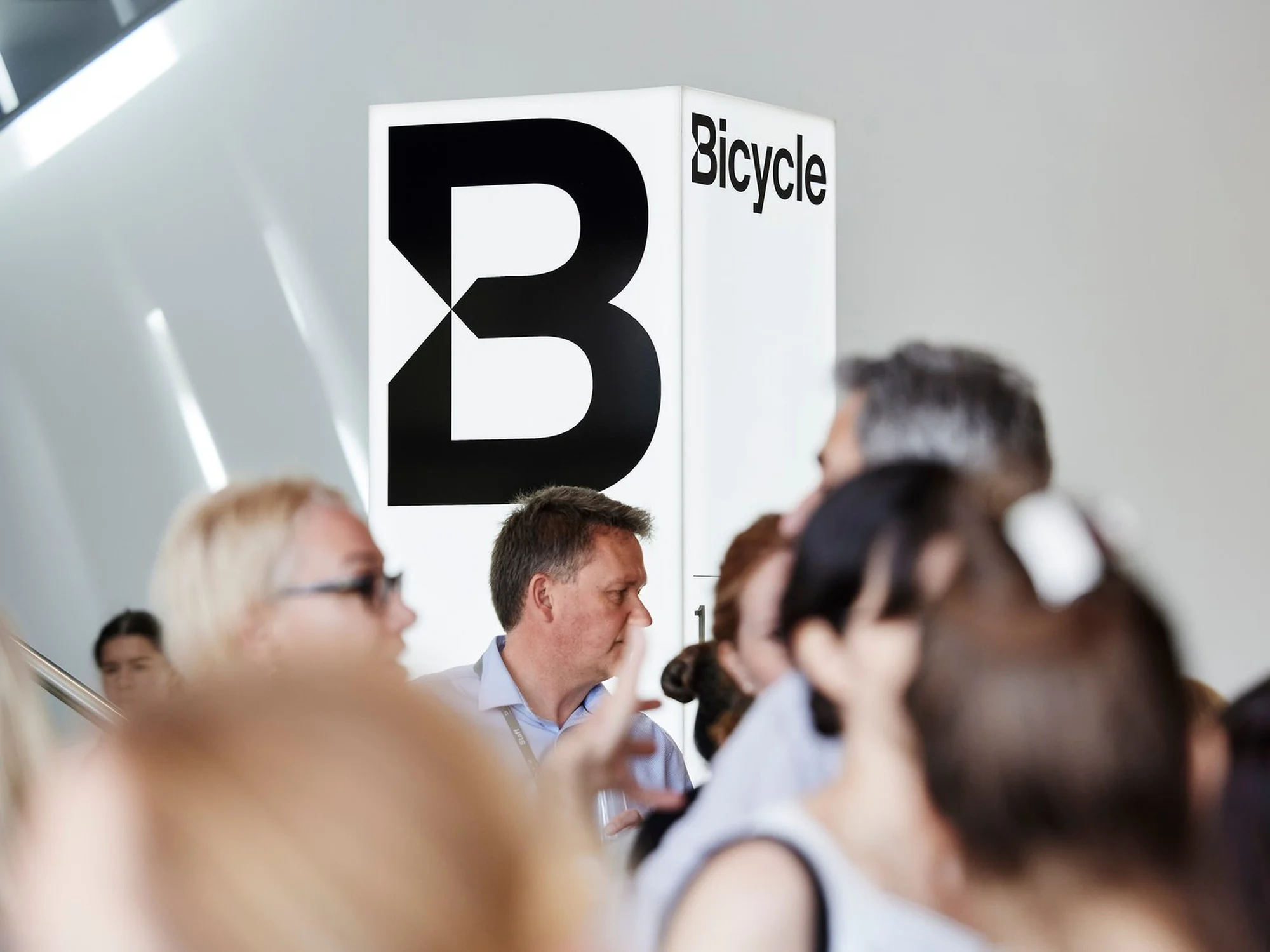 A group of people are gathered indoors near a sign with a large black 'B' and the word 'Bicycle' at what appears to be an event.