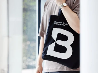 Person wearing a grey t-shirt holding a black tote bag with white typography and a large letter B logo.
