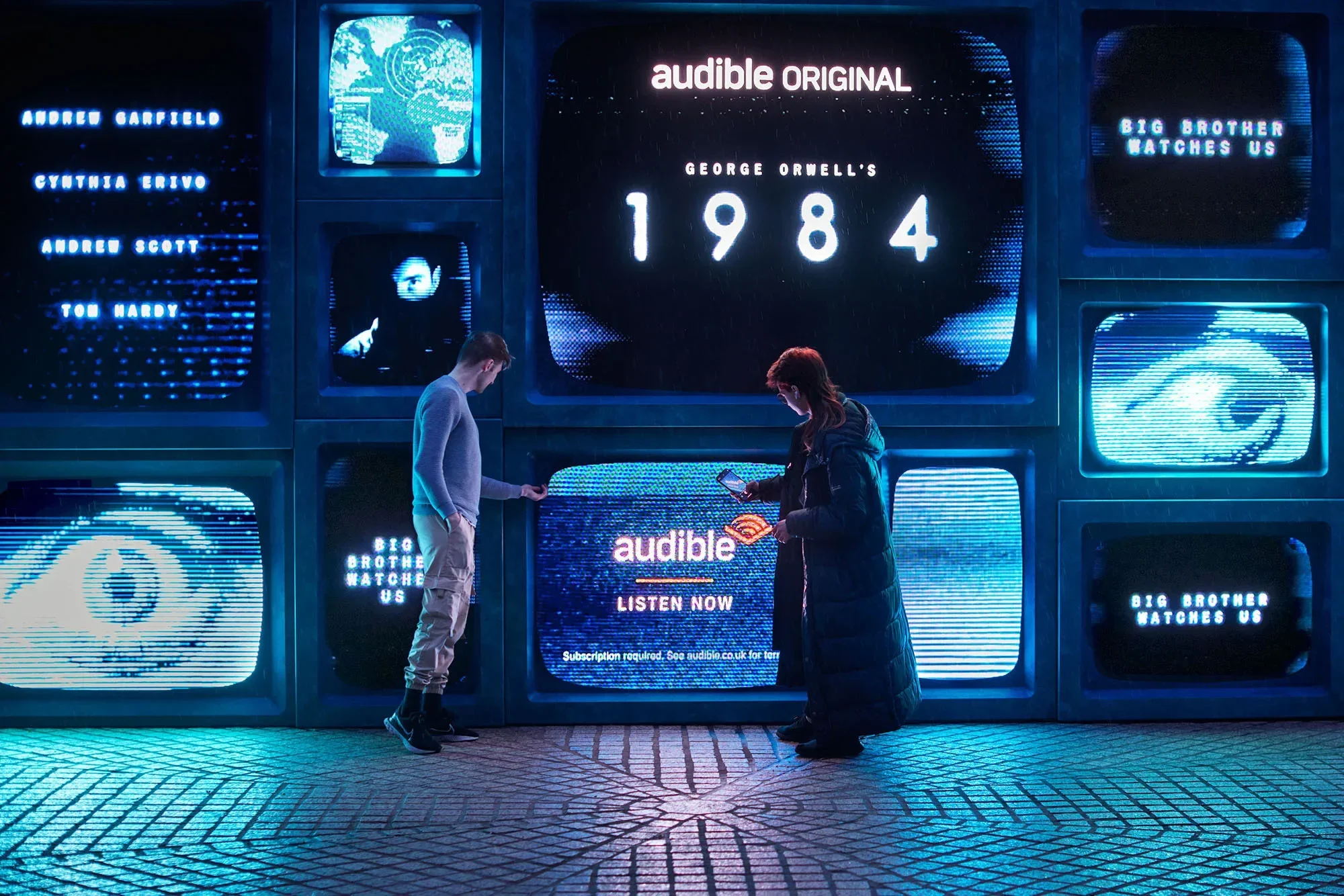 Two adults are viewing a display of stacked monitors that show the cover and promotional material for "1984" by George Orwell, an Audible Original.