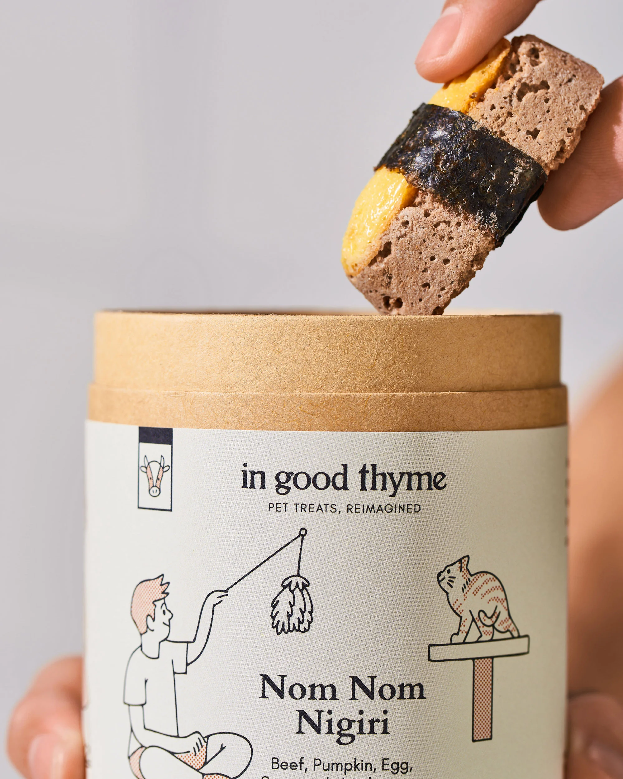 Hand holding a dog treat shaped like nigiri sushi, placed above the container. Dog treats container shows illustrations of a cat and human.