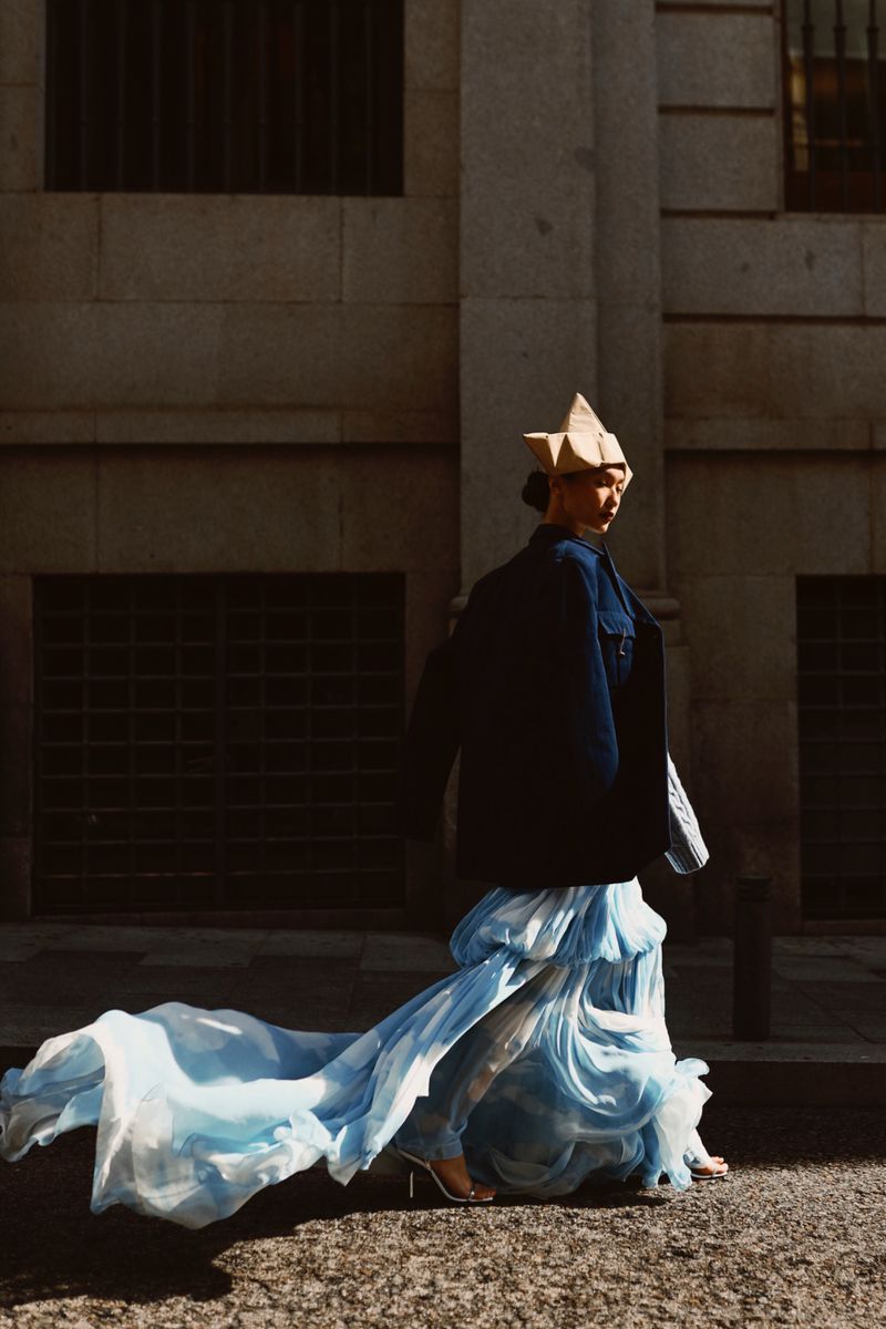 Fashionable adult walking outdoors wearing an architectural paper hat, dark jacket, and flowing blue dress with metallic high heels.