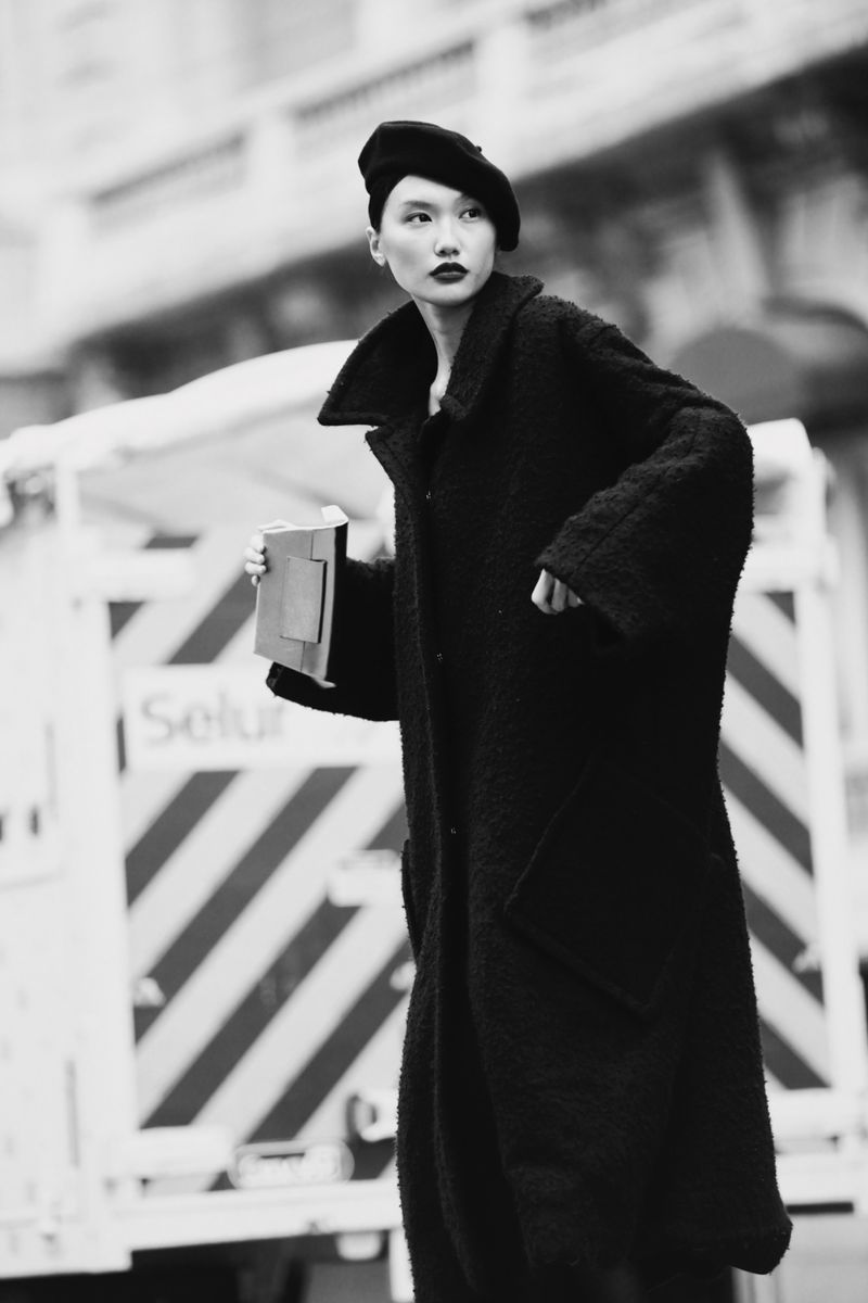 Monochrome outdoor fashion shot of an Asian woman in a black beret and long coat, holding a portfolio against a blurred street backdrop.