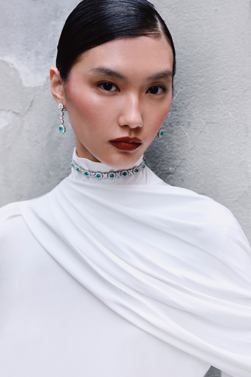Close-up of an elegant Asian woman wearing a white draped dress and a stunning emerald and diamond necklace and earrings against a grey wall.
