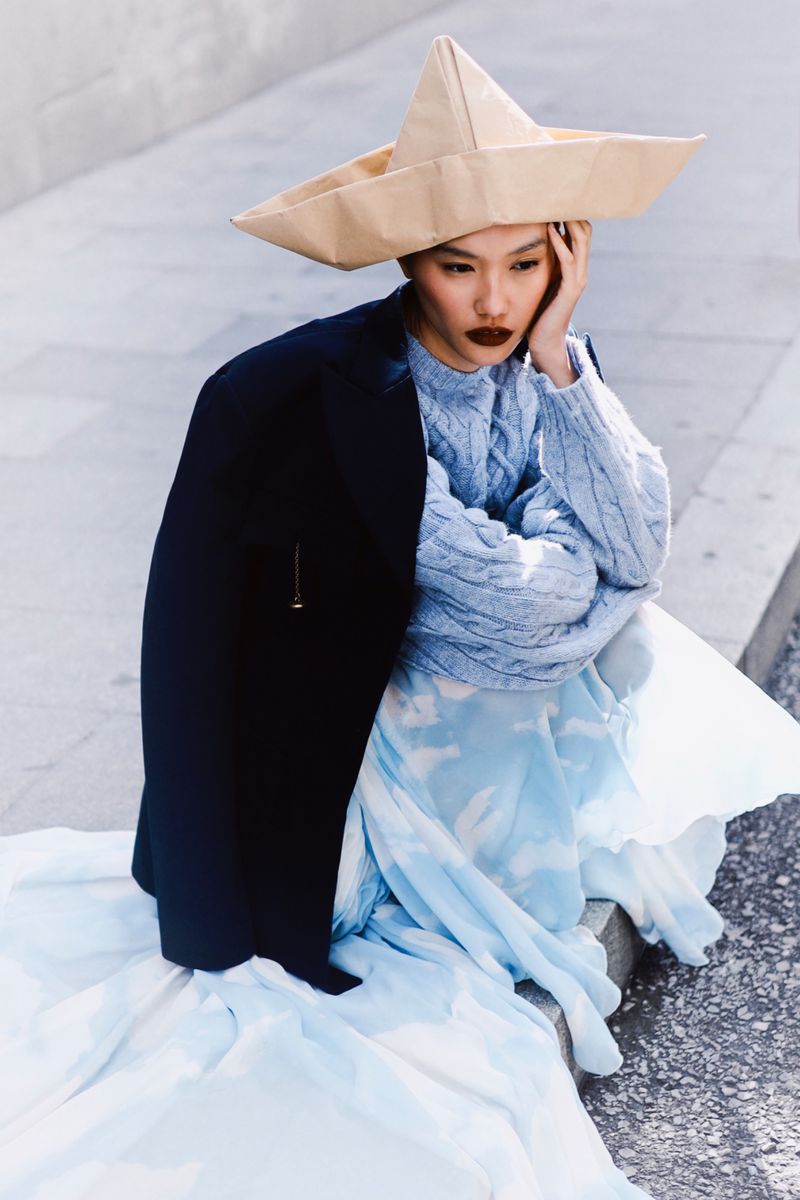 A woman wearing a paper hat and blue dress is seated on concrete, posed outdoors in editorial fashion photography with overcast lighting.