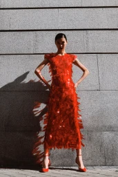 Model in a red fringe dress with hands on hips standing against a textured stone wall in bright sunlight