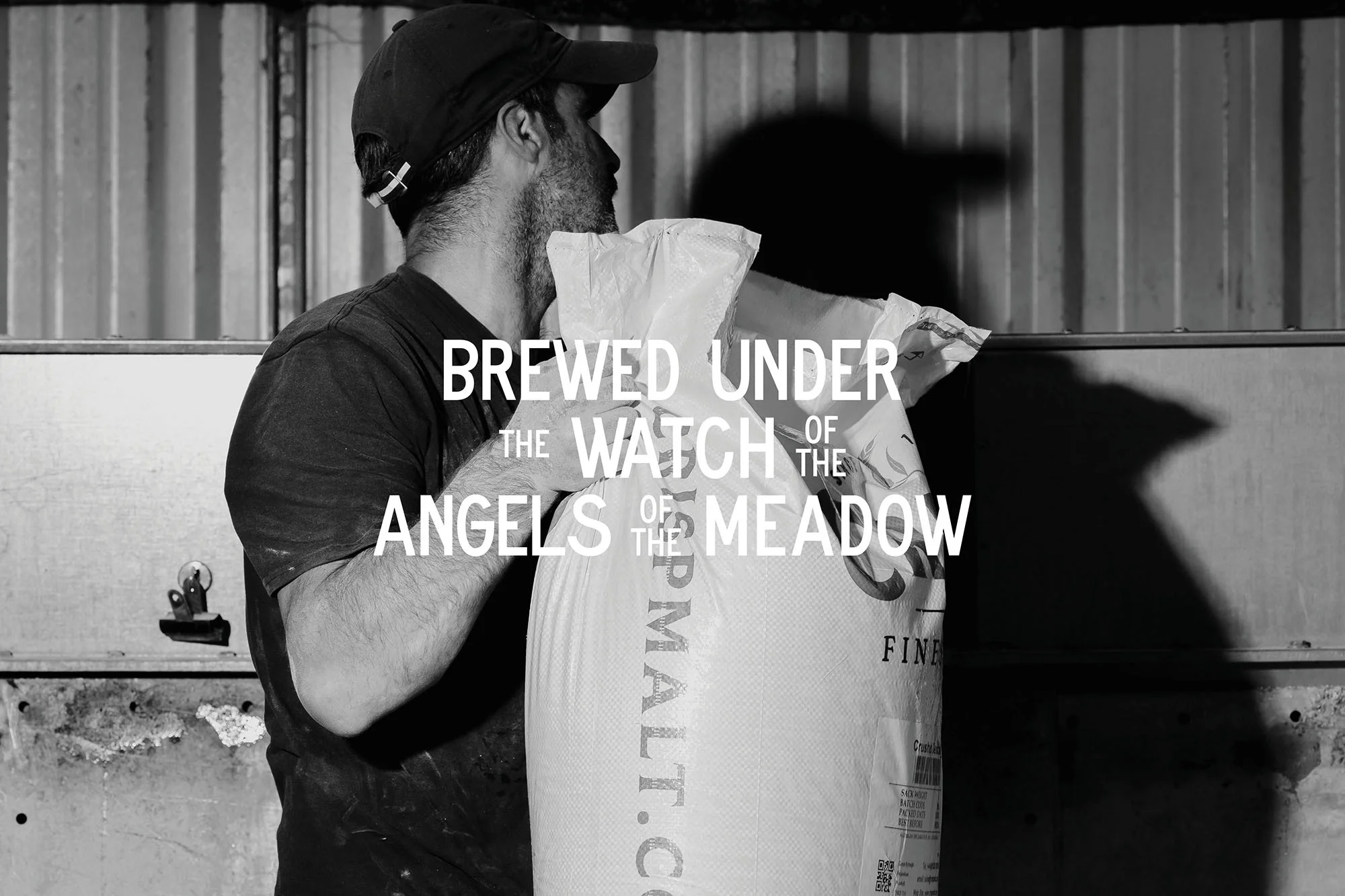 Monochrome image of a man in a cap carrying a large bag with text. The scene is set against a metal wall.