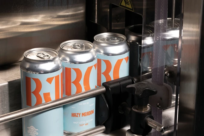Several cans of light beer move along a mechanical conveyor inside of a packaging machine, in a bright, industrial-style setting.