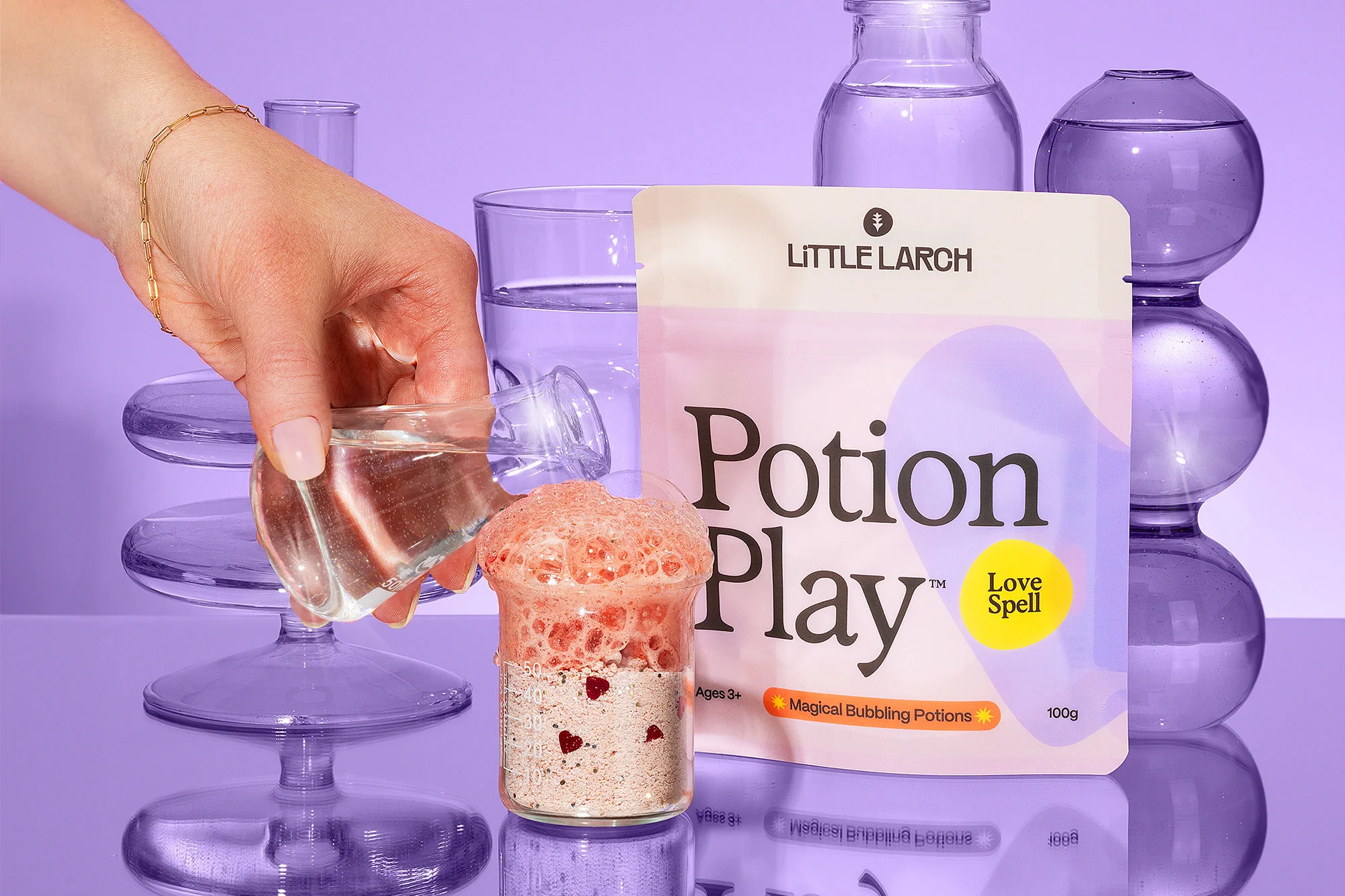 Woman's hand pours water into a glass of bubbling potion next to a Little Larch Potion Play packet with laboratory glassware.
