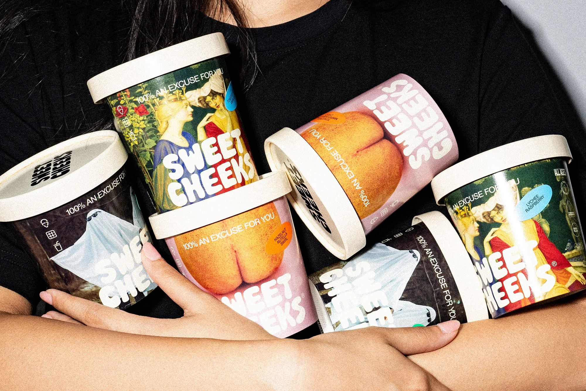 A woman holds several tubs of 'Sweet Cheeks' ice cream with artistic labels, including peaches, classical art, and a ghost.