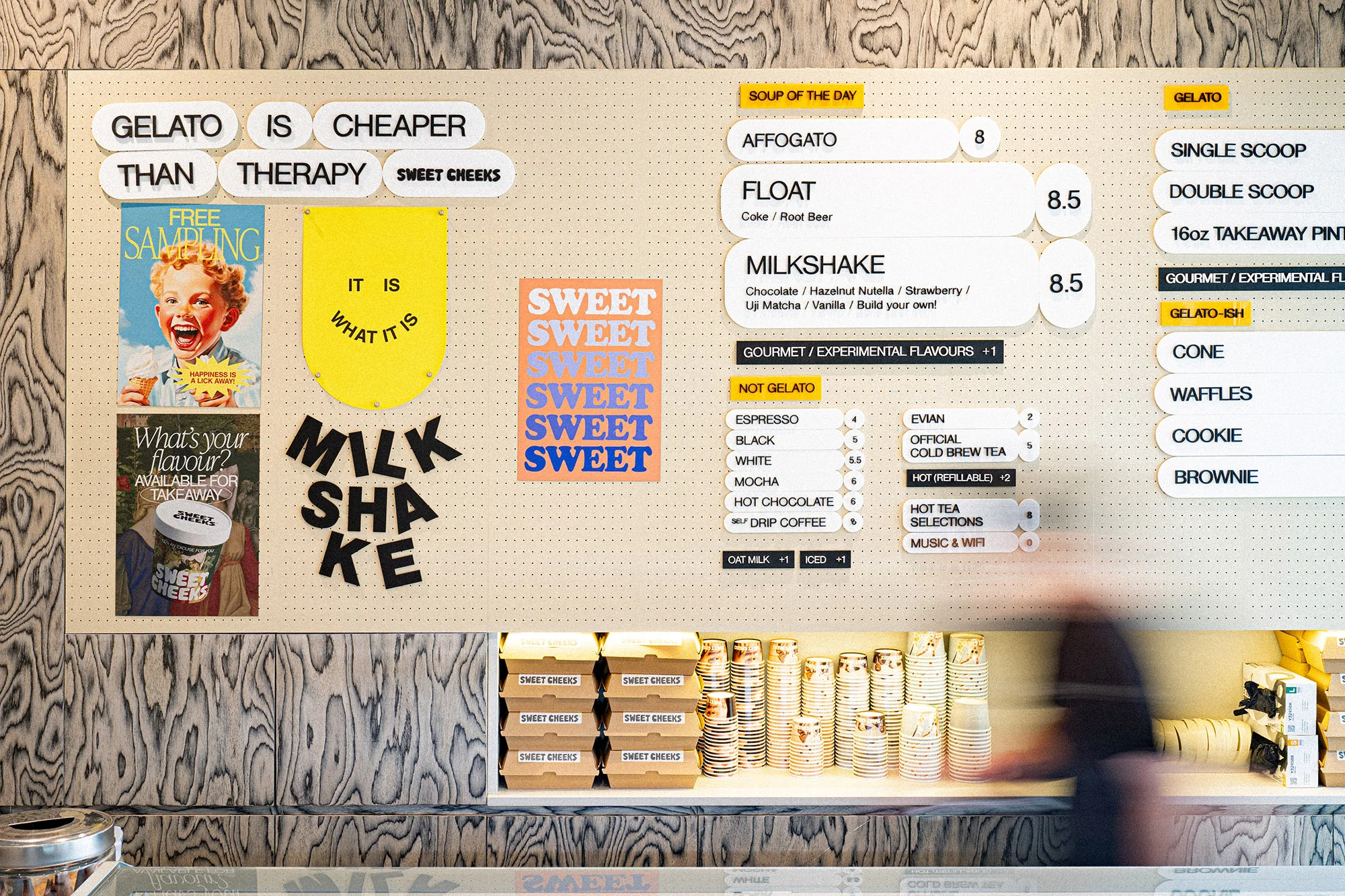 Menu board with food and beverage options, including gelato, milkshakes, coffee, tea, and desserts, alongside promotional posters and signage.
